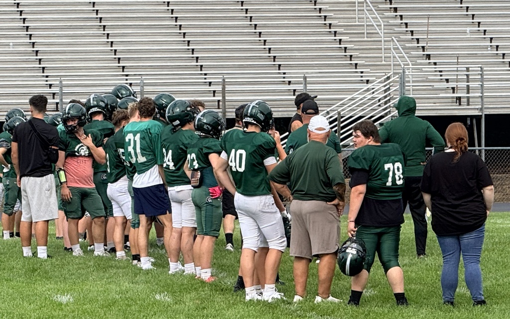 Hampshire High School football players and coaches gather along the field. WVSDB student #76 stands with the team in a green football jersey and helmet.