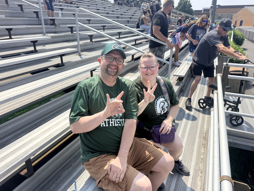 Staff members sit together in the bleachers at a Hampshire High School football event, smiling at the camera and signing “78.” Both wear green Hampshire athletics apparel.