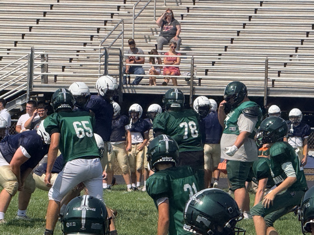 Hampshire High School football players gather on the field during a practice or scrimmage. WVSDB student #76 stands among his teammates wearing a green jersey and football helmet.