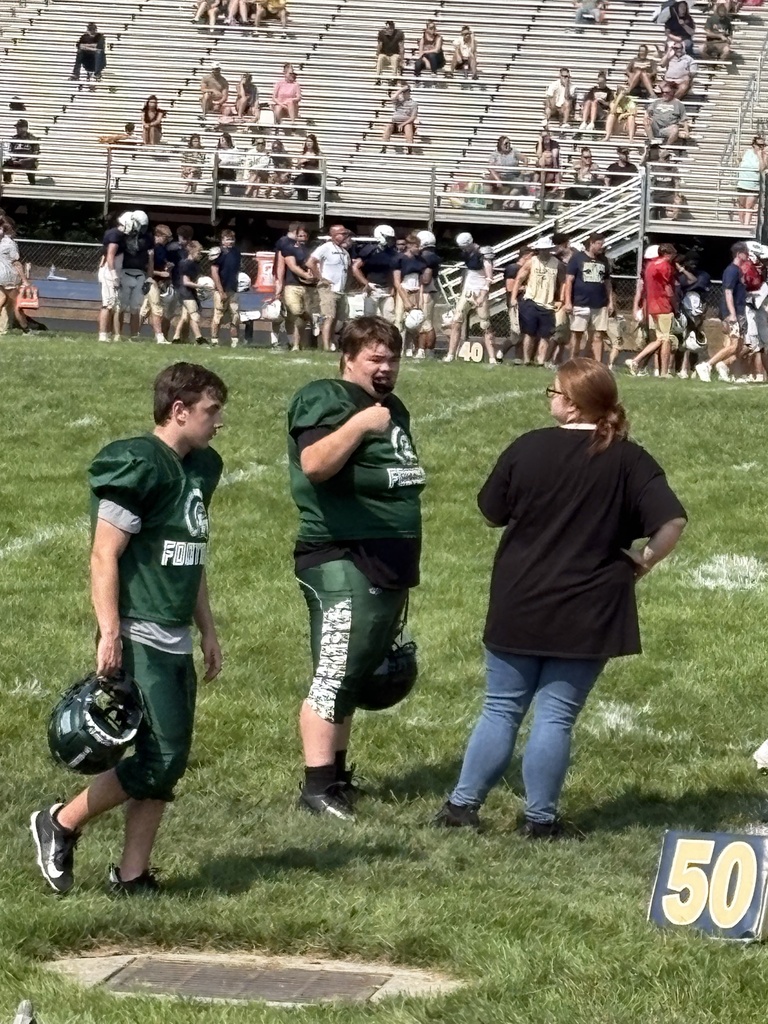 WVSDB student #76 stands on the football field in his green Hampshire High School uniform while talking with a staff member. Other players and spectators are visible in the background.