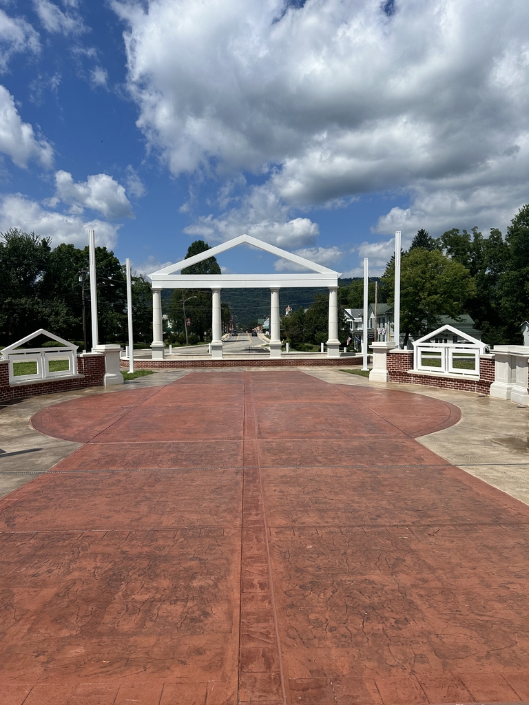 Wide view of the completed WVSDB Administration Building memorial, featuring the white columned structure, brick walls, flagpoles, and a large red walkway leading toward the memorial beneath a blue, partly cloudy sky.