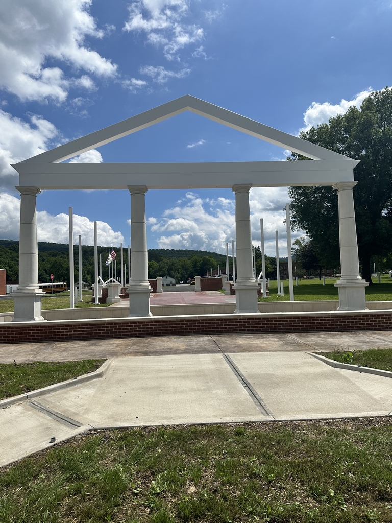 Close view of the WVSDB Administration Building memorial, featuring four white columns and a peaked structure overlooking the memorial plaza, flagpoles, and campus beneath a bright blue sky.