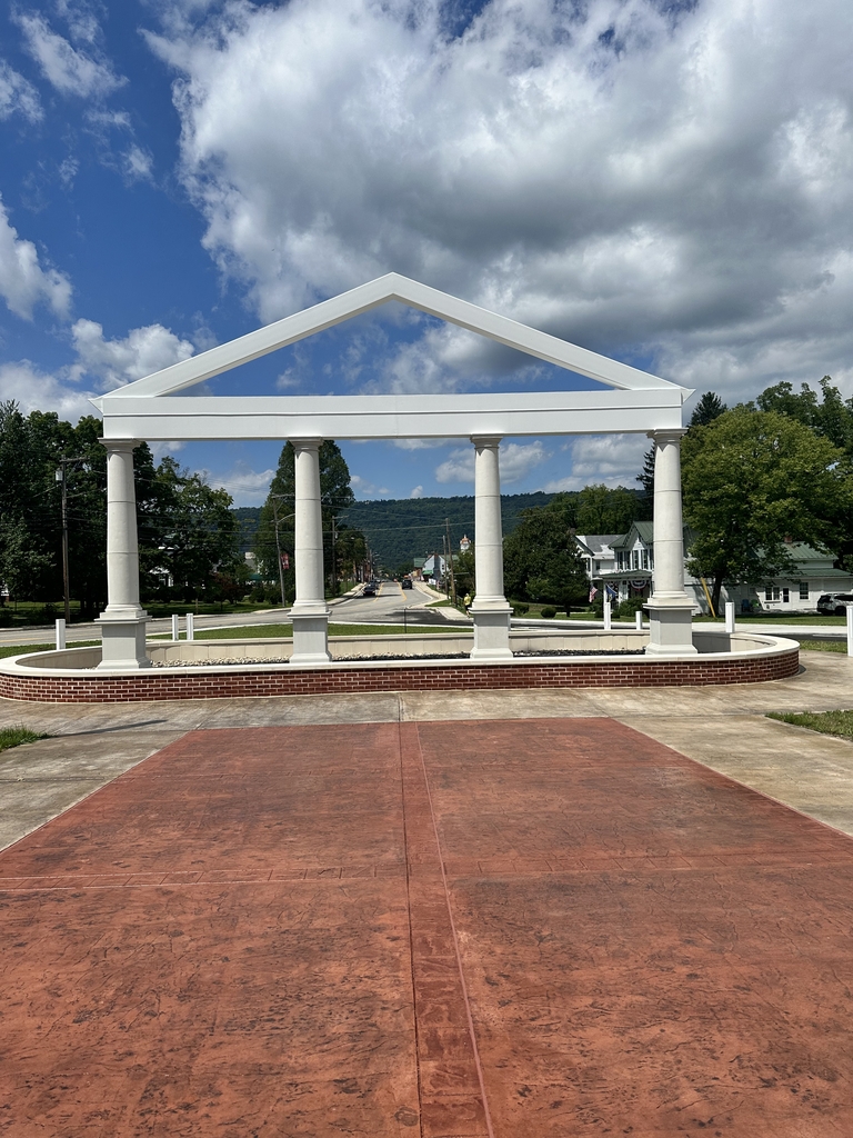 Front view of the WVSDB Administration Building memorial, featuring four white columns and a peaked structure with the town of Romney and mountains visible in the background beneath a partly cloudy sky.
