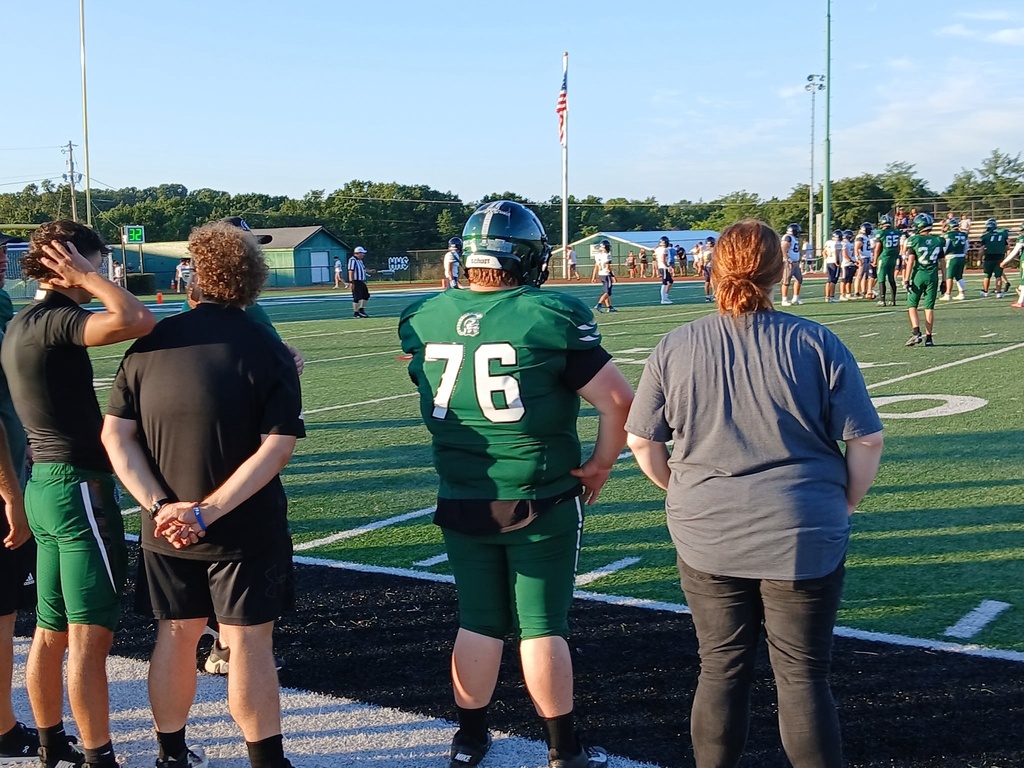 A football player wearing a green number 76 jersey and helmet stands on the sideline watching the action on the field during a scrimmage. Other players, coaches, and spectators are visible across the field on a sunny evening.