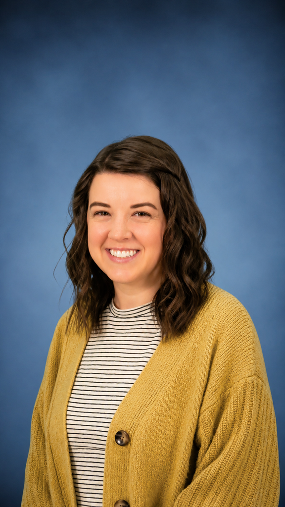Professional portrait of a smiling woman with shoulder-length wavy brown hair, wearing a mustard yellow cardigan over a black-and-white striped shirt, posed against a soft blue background.