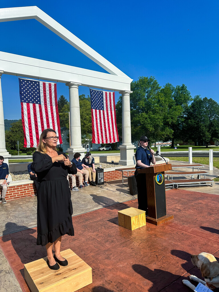 WVSDB student speaks at the podium while an interpreter signs beside them, with the memorial and American flags in the background.