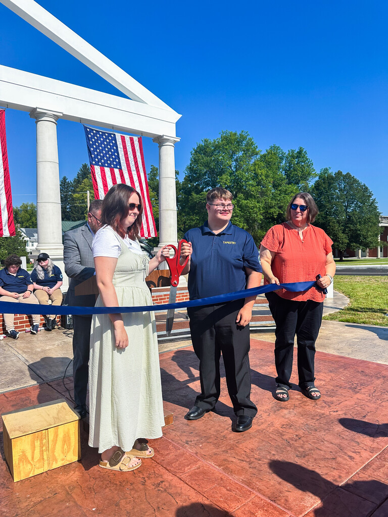 WVSDB student holds ceremonial scissors at a blue ribbon during the Administration Building Memorial ribbon-cutting ceremony, surrounded by staff.