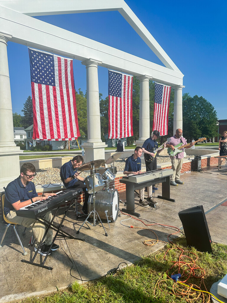 WVSDB student musicians perform on keyboards, drums, guitar, and bass in front of the Administration Building Memorial, decorated with large American flags.