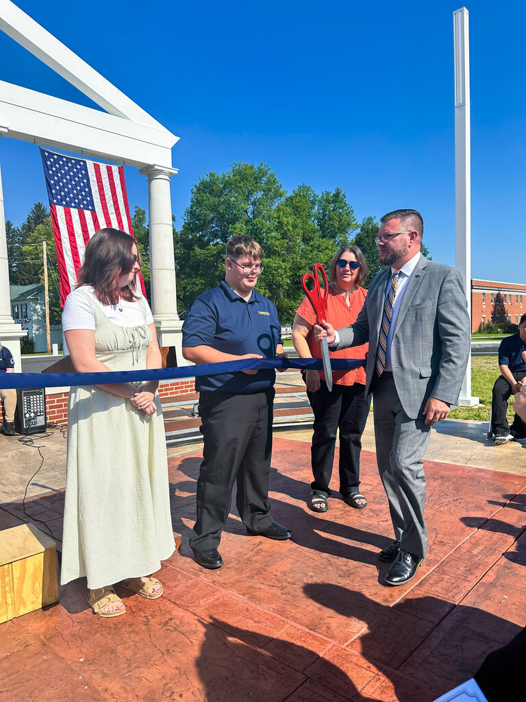 WVSDB student and school representative hold oversized ceremonial scissors at the blue ribbon during the Administration Building Memorial dedication.