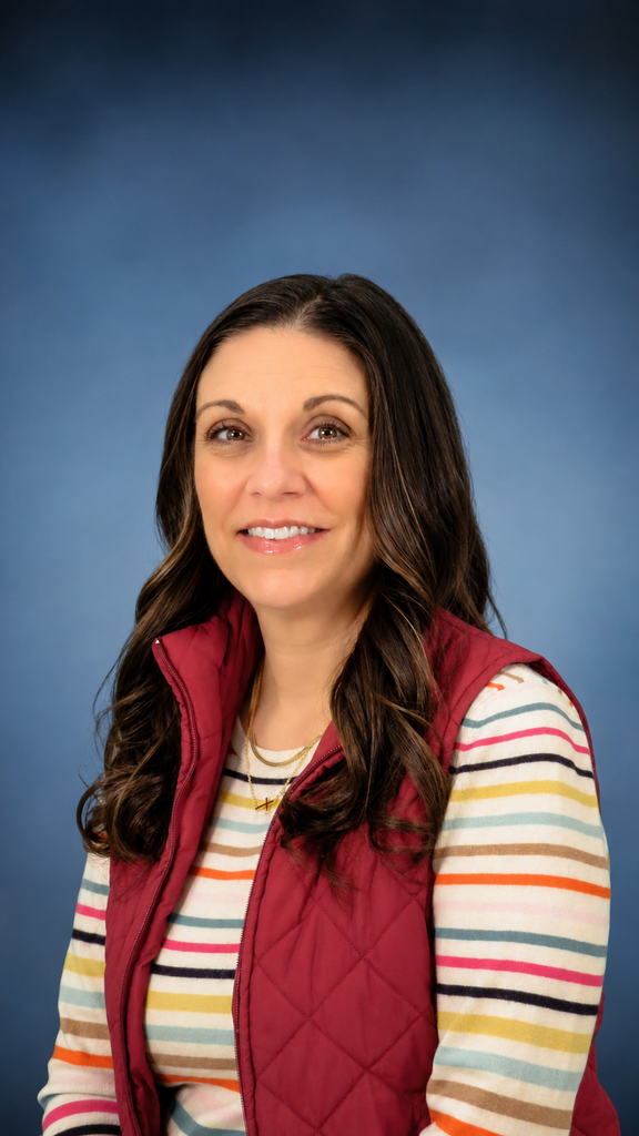 Professional portrait of a smiling woman with long dark brown hair, wearing a red quilted vest over a multicolored striped shirt, posed against a soft blue background.