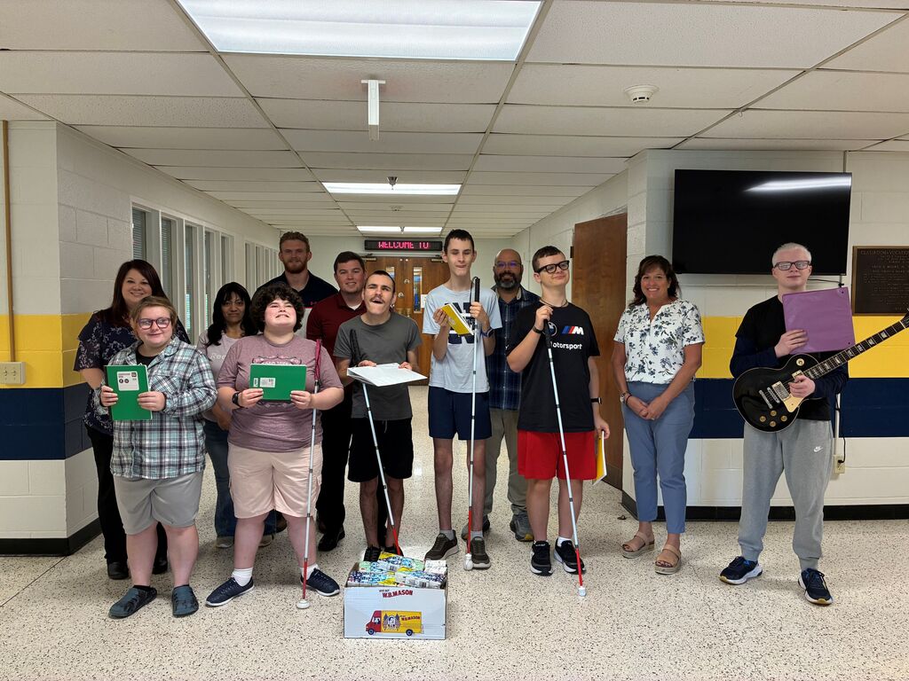 WVSDB students and staff gather in a school hallway with donated school supplies from the Bank of Romney. Several students hold notebooks and folders while posing together with staff.