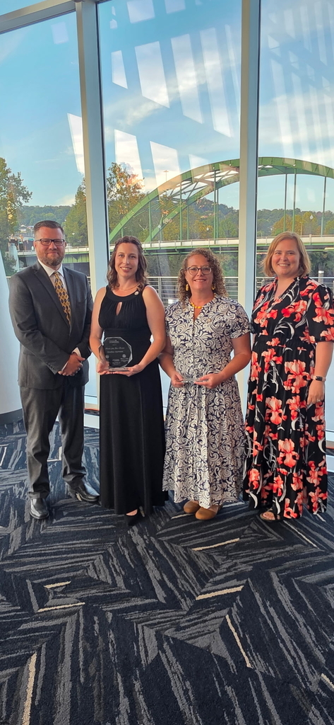Ms. Heather Hartman and Mrs. Joanna Gilbert stand together holding their finalist awards alongside two other attendees at the West Virginia Teacher and Service Personnel of the Year celebration. A large window behind the group overlooks a green bridge and surrounding landscape.