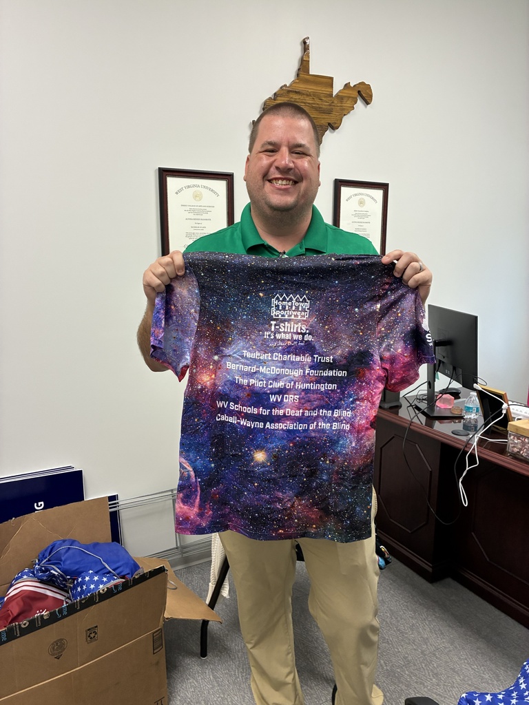 A smiling man in a green Cabell-Wayne Association of the Blind polo holds up a galaxy-print SCIVIS T-shirt featuring sponsor and organization names, including WVSDB.