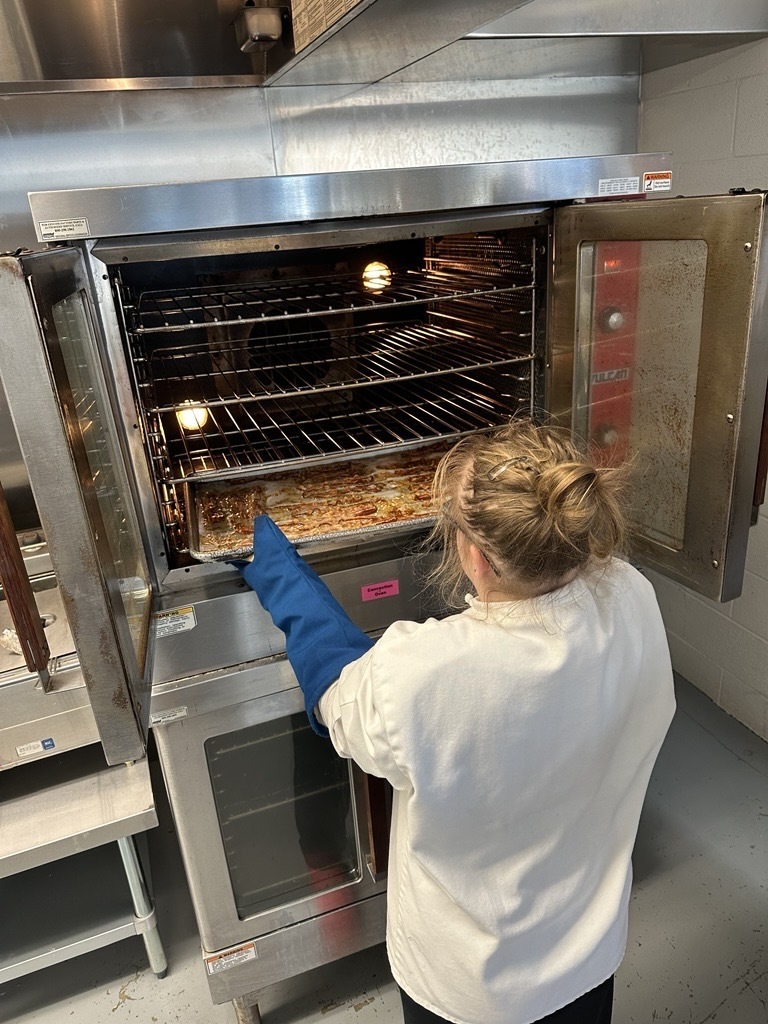 Student wearing an oven mitt removes a tray of cooked bacon from a commercial oven in the Appalachia Provisions kitchen.