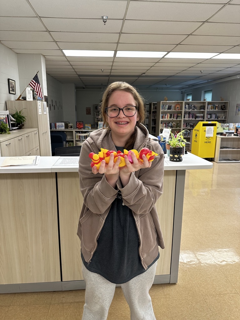 A smiling student stands in the school library holding several colorful “I Love You” ducks she collected.