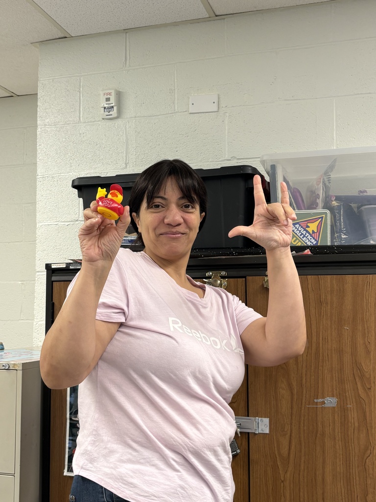 A staff member smiles while holding an “I Love You” duck and making the ASL “I love you” handshape.