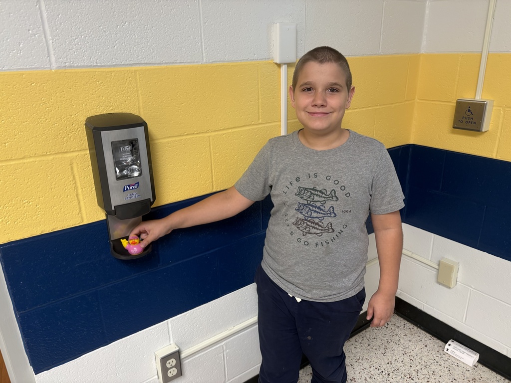 A smiling student stands beside a wall-mounted hand sanitizer dispenser, where he discovered an “I Love You” duck.
