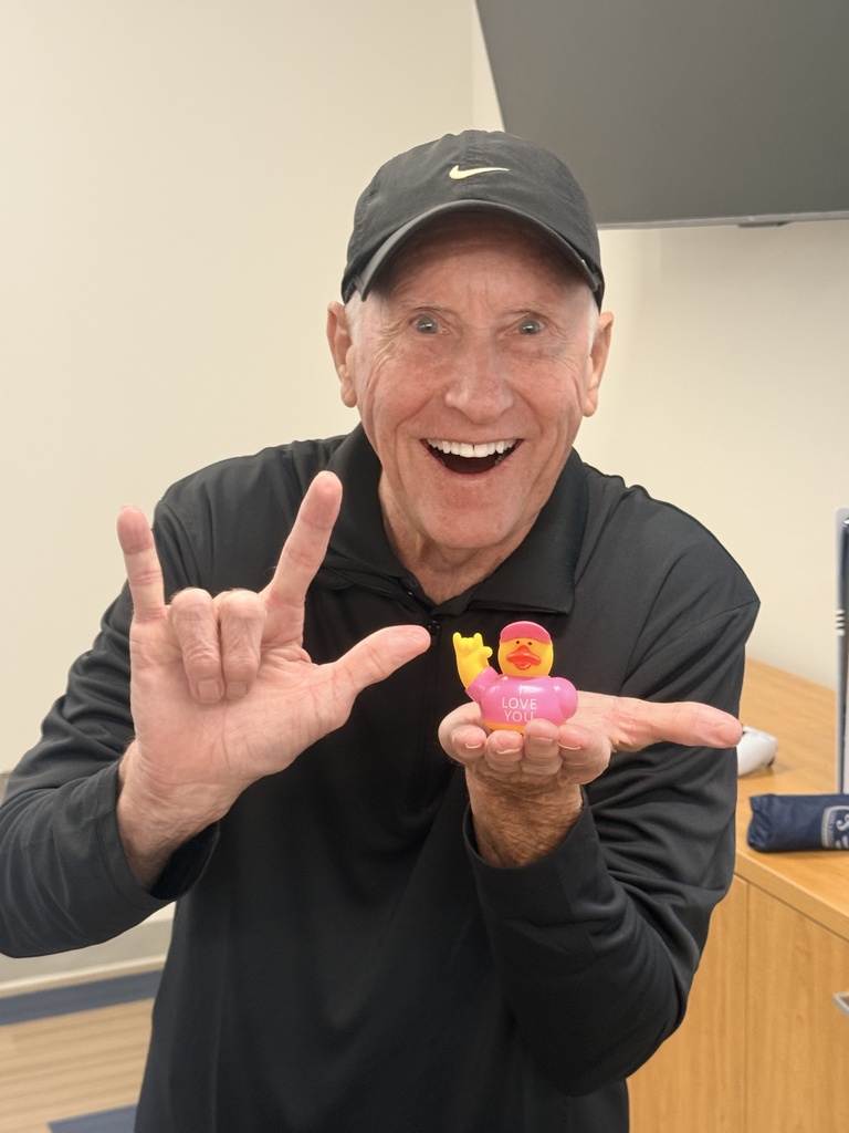 A smiling visitor holds an “I Love You” duck while making the ASL “I love you” handshape with his other hand.