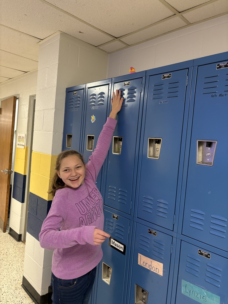 A smiling student reaches toward an “I Love You” duck hidden high on top of the blue school lockers.