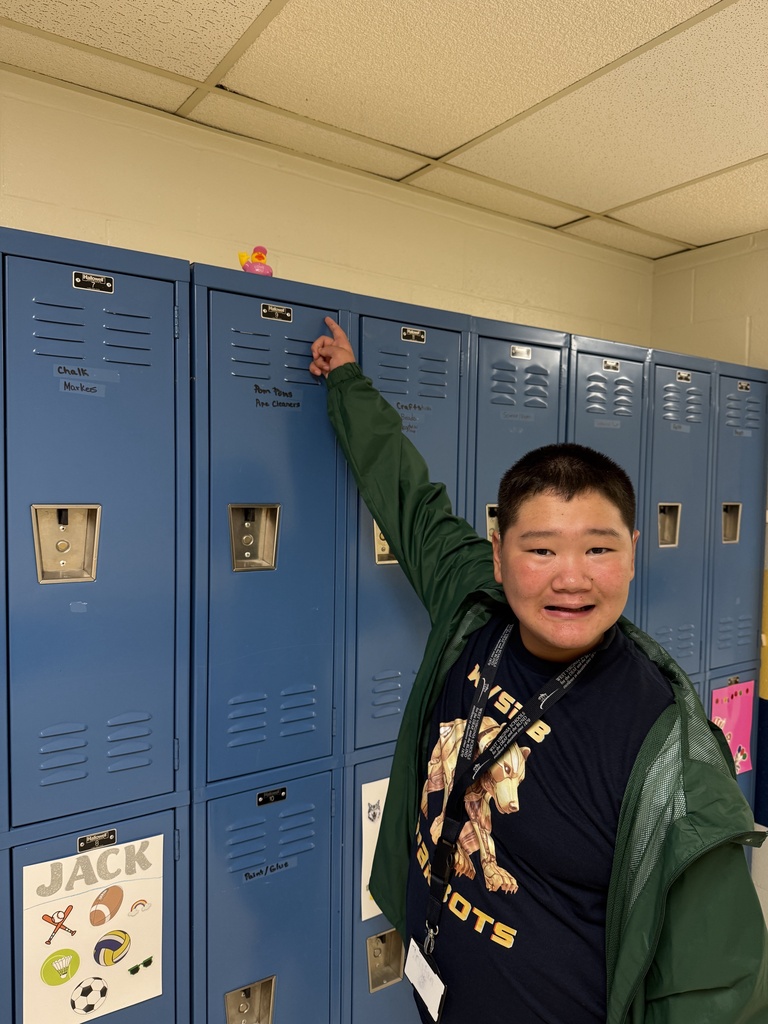 A smiling student points to an “I Love You” duck hidden on top of a row of blue lockers.