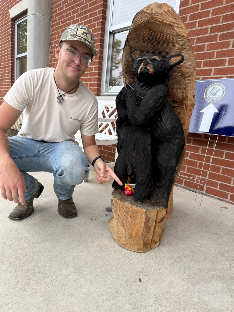 A student crouches beside a carved wooden black bear, pointing to an “I Love You” duck hidden at the bear’s feet.