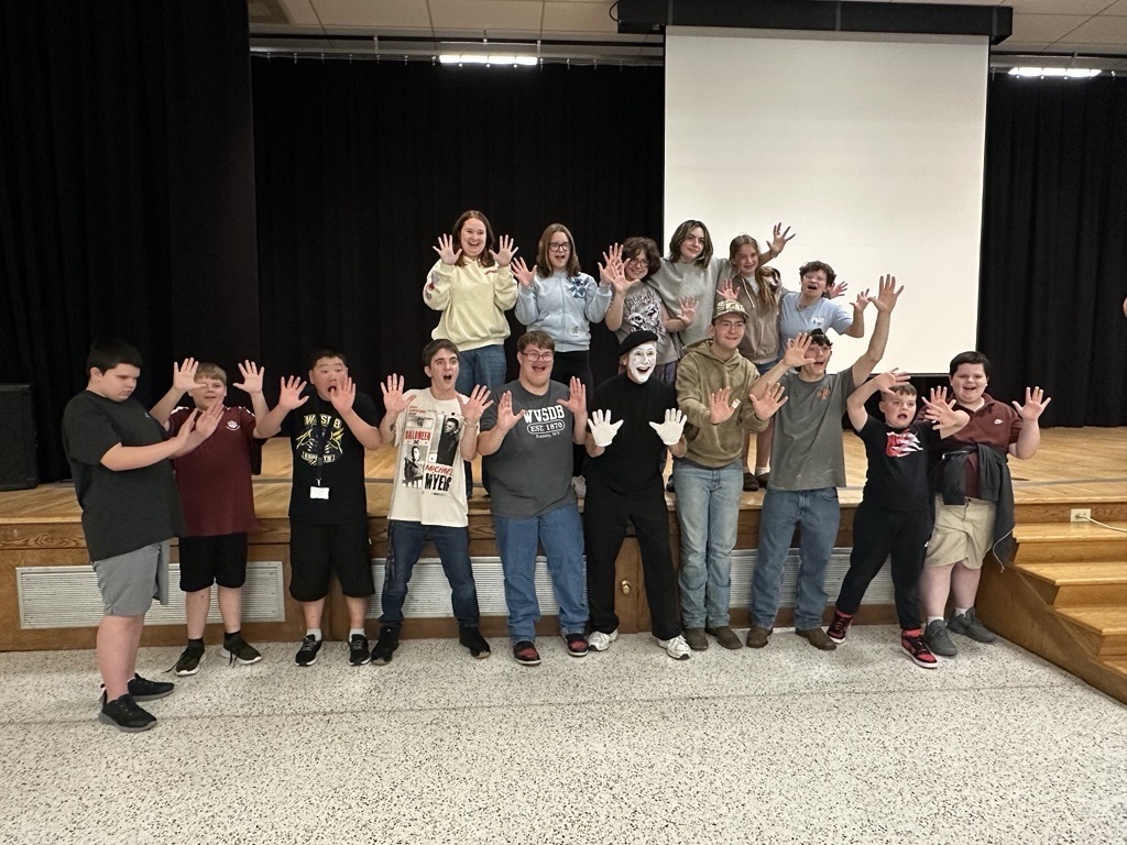 JJ Jones poses in the center of a large group of WVSDB students after his performance, with students smiling and holding their hands up toward the camera.