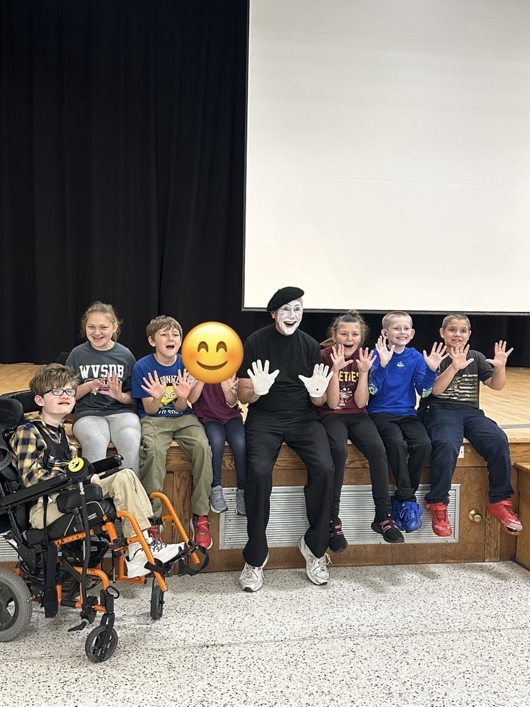 JJ Jones sits with a group of younger WVSDB students at the edge of the stage as they smile and hold up their hands for a group photo.