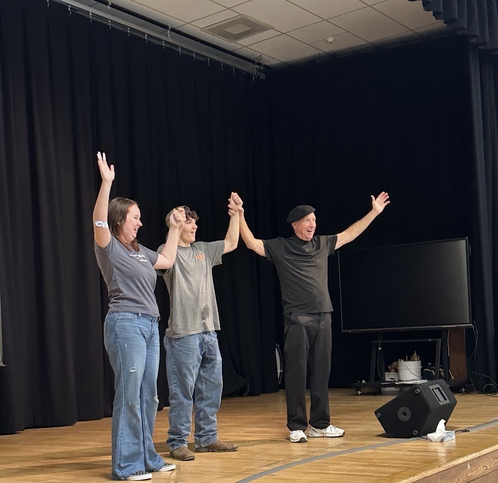 Deaf Mime JJ Jones stands on stage with two WVSDB participants, holding their hands up after performing together.