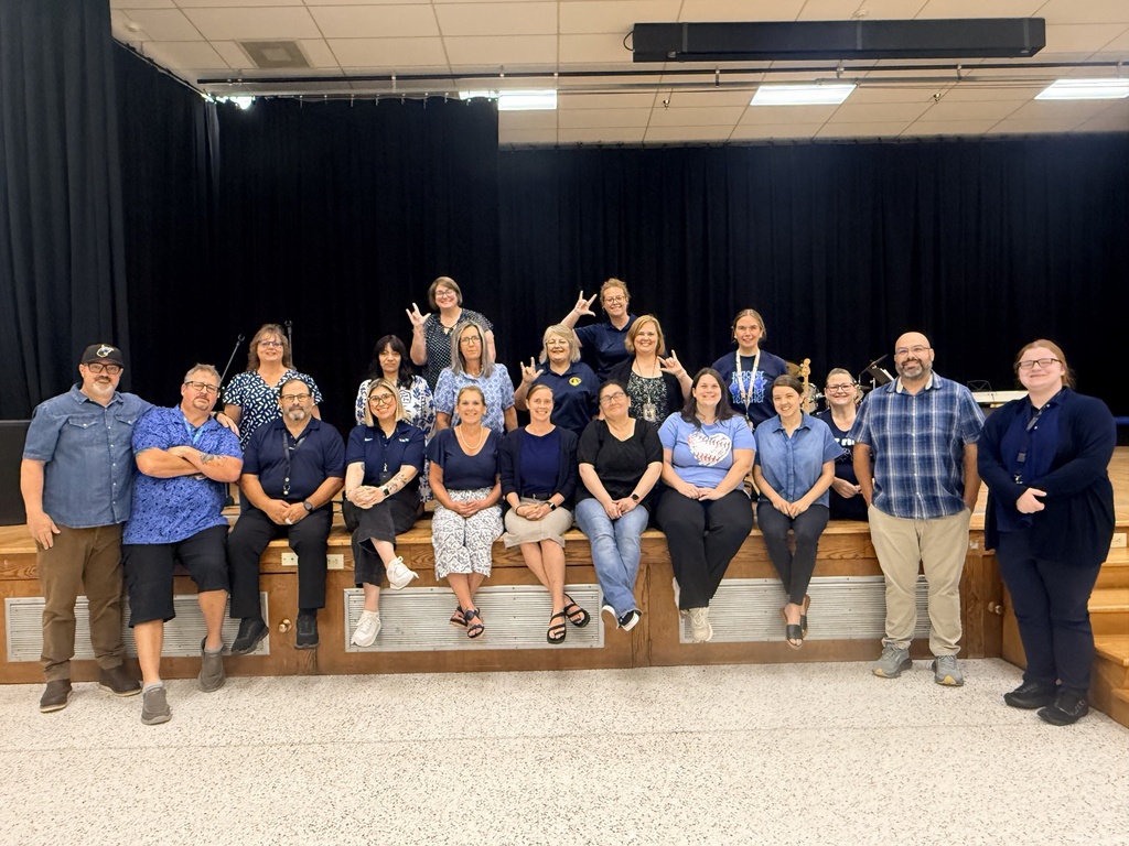 Group photo of WVSDB staff members gathered in front of a dark stage curtain. Staff are wearing various shades of blue in recognition of Deaf Awareness Month, and several people are making the ASL “I love you” handshape.