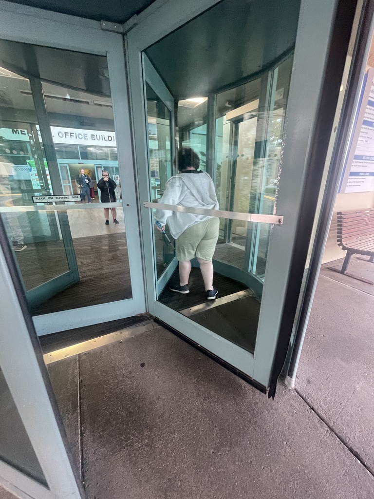 A WVSDB Blind/Low Vision student continues navigating through a revolving door using a white mobility cane during community-based O&M instruction.