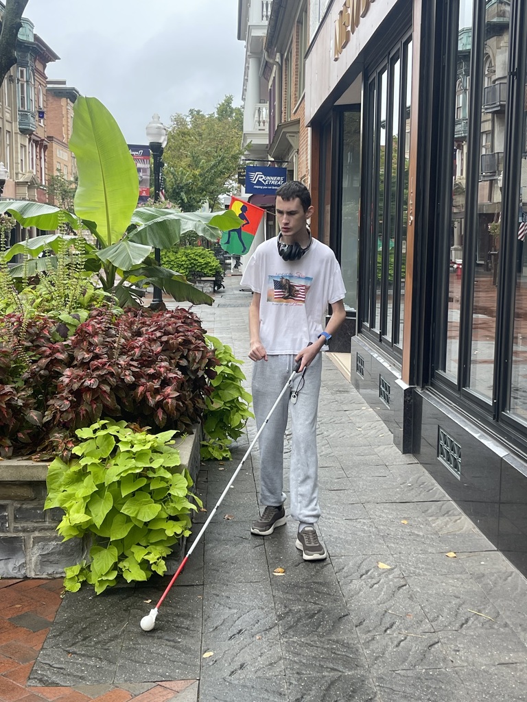 A WVSDB Blind/Low Vision student travels along a downtown Winchester sidewalk using a white mobility cane during community-based Orientation & Mobility instruction.