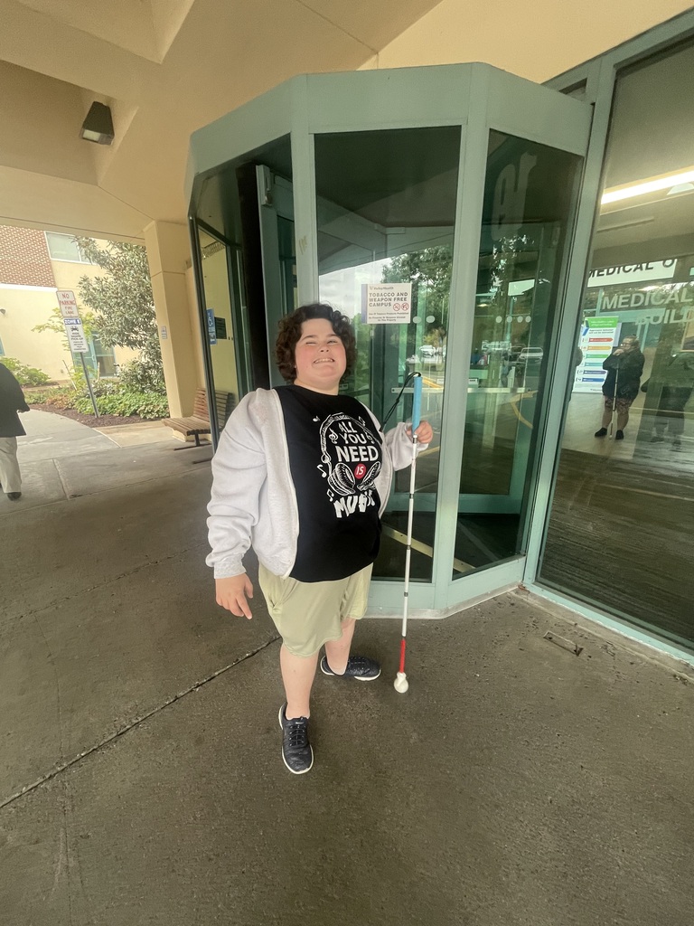 A WVSDB Blind/Low Vision student smiles while standing outside a building in Winchester, Virginia, holding a white mobility cane.