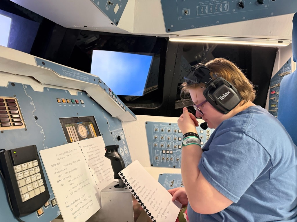 WVSDB student wearing a headset sits at the controls of a spacecraft simulator while following printed instructions.