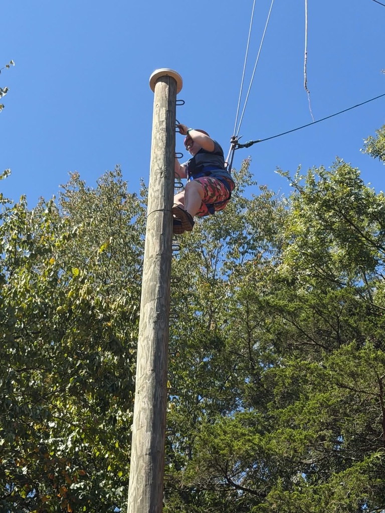 WVSDB student wearing a safety harness climbs a tall wooden pole as part of an outdoor challenge course.