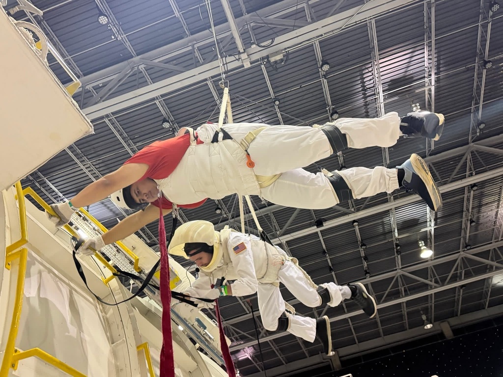 Two participants in astronaut training suits are suspended horizontally while completing a simulated space mission activity.