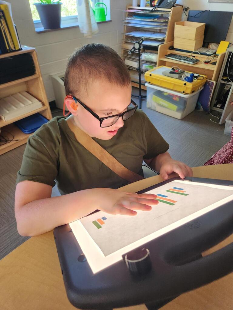 A student wearing glasses examines a bar graph displayed on an illuminated light box. The student places a hand on the graph while reviewing data collected during the stomp rocket science experiment.