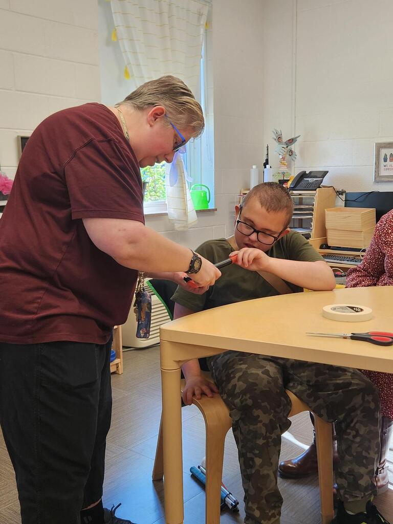 A teacher assists a student wearing glasses with preparing a stomp rocket during a hands-on science activity. The student holds part of the rocket assembly while the teacher helps adjust and secure the components.