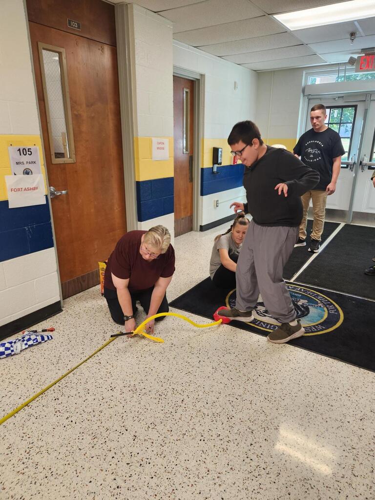 A student steps onto the air pump of a stomp rocket launcher in a school hallway. A teacher kneels nearby to position the launcher while classmates and staff observe the experiment.