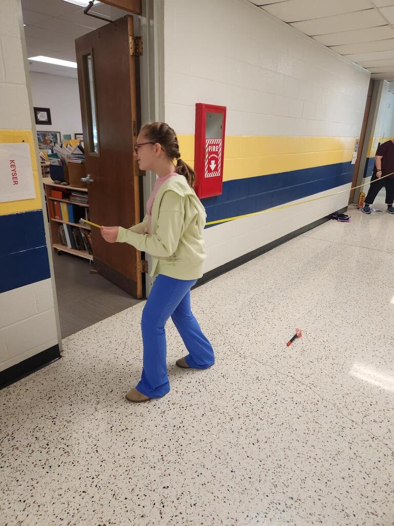A student uses a measuring tape stretched along the school hallway to measure the distance traveled by a stomp rocket. A small rocket rests on the floor nearby as the student records the results of the experiment.