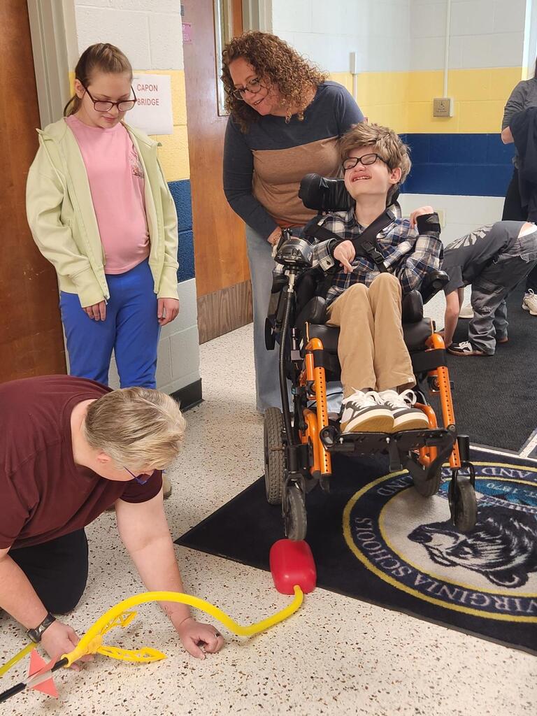 A smiling student using a wheelchair participates in a stomp rocket experiment alongside classmates and teachers. One teacher kneels to prepare the launcher while another stands beside the student, providing support during the activity.