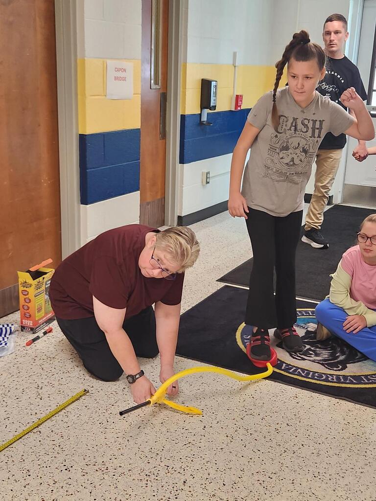 A student steps onto the air pump of a stomp rocket launcher while a teacher kneels to position the rocket. Classmates watch nearby as the student participates in the hands-on World Space Week science experiment.