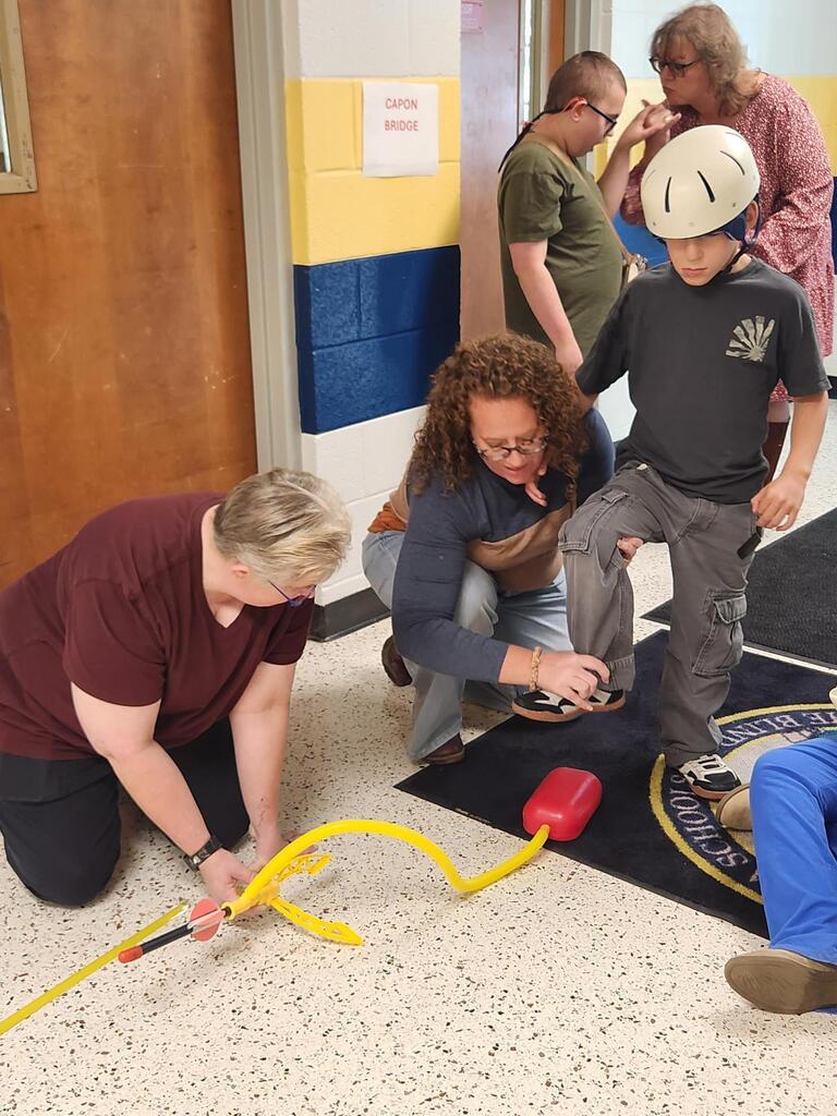 A student wearing a protective helmet prepares to activate a stomp rocket launcher by stepping onto its air pump. Two teachers kneel nearby to position the rocket and assist the student while classmates observe.