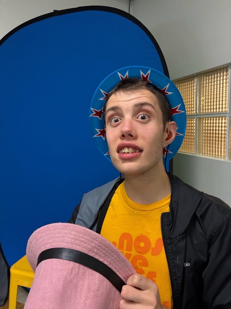 A young adult makes a playful expression while wearing a colorful novelty hat and holding a pink hat in front of a blue photo booth backdrop.