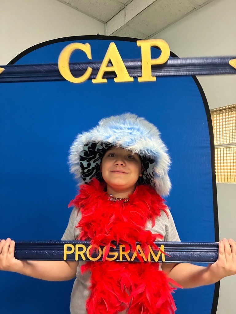A student wearing a fluffy blue hat and bright red feather boa holds a large decorative CAP Program photo frame in front of a blue backdrop.