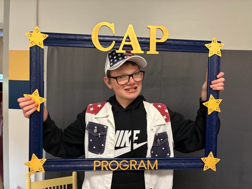 A smiling student wearing glasses, a patriotic cap, and a red, white, and blue vest holds a decorative CAP Program photo frame.