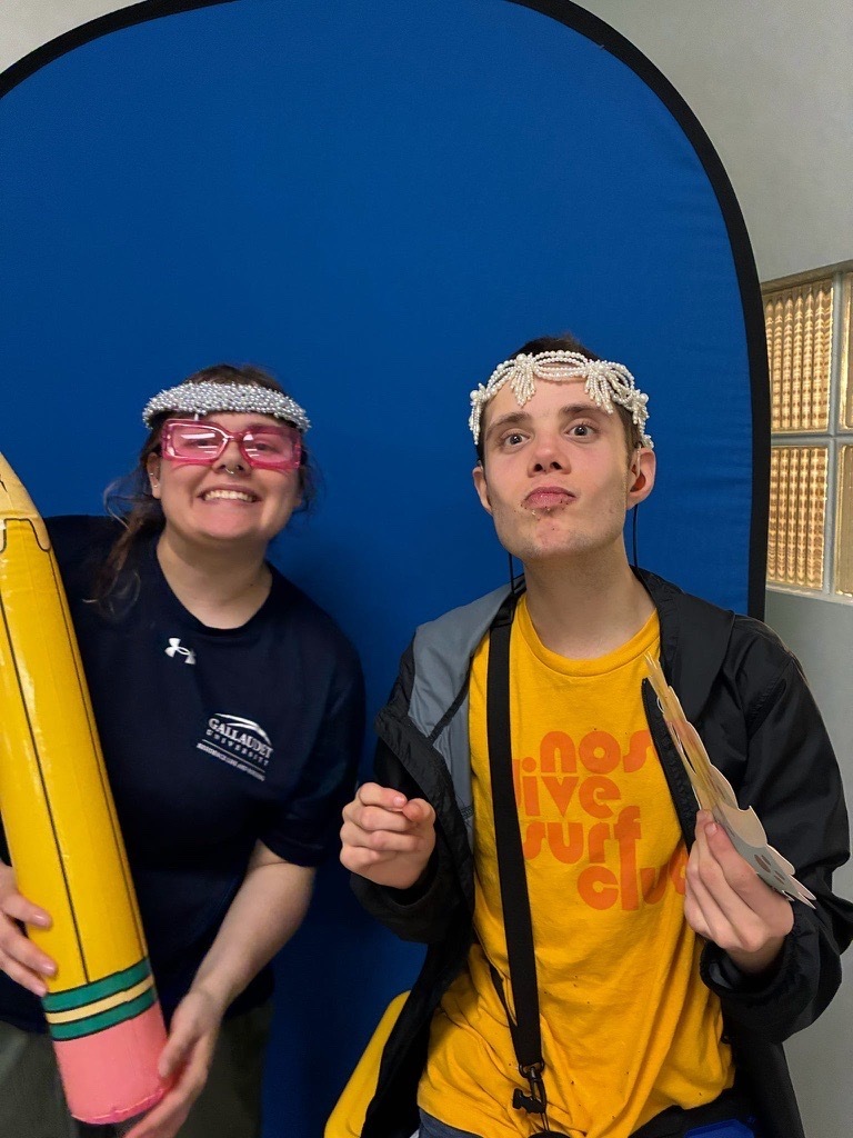 Two smiling young adults pose with playful accessories, including pink glasses, pearl headpieces, and an oversized pencil prop.