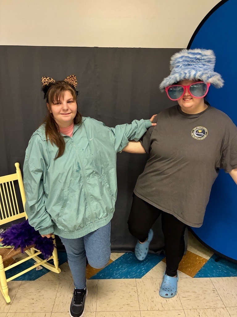 Two individuals pose together wearing leopard-print ears, oversized pink sunglasses, and a fluffy blue hat in the photo booth area.