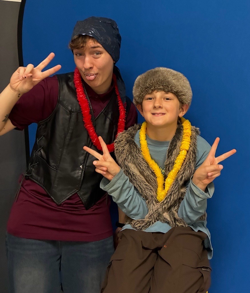 Two individuals pose with peace signs while wearing furry hats, colorful leis, and a black vest in front of a blue photo booth backdrop.