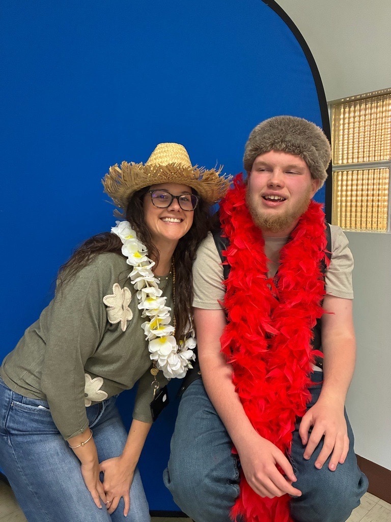 Two smiling individuals pose in front of a blue backdrop wearing playful photo booth accessories, including a straw hat, furry hat, and red and white leis.