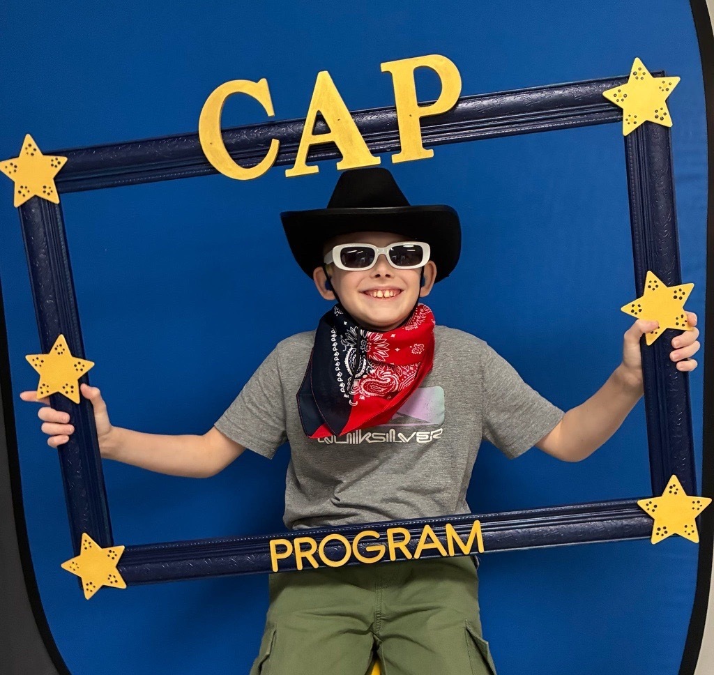 A smiling student wearing a cowboy hat, white sunglasses, and red bandana poses inside a decorative CAP Program photo frame.